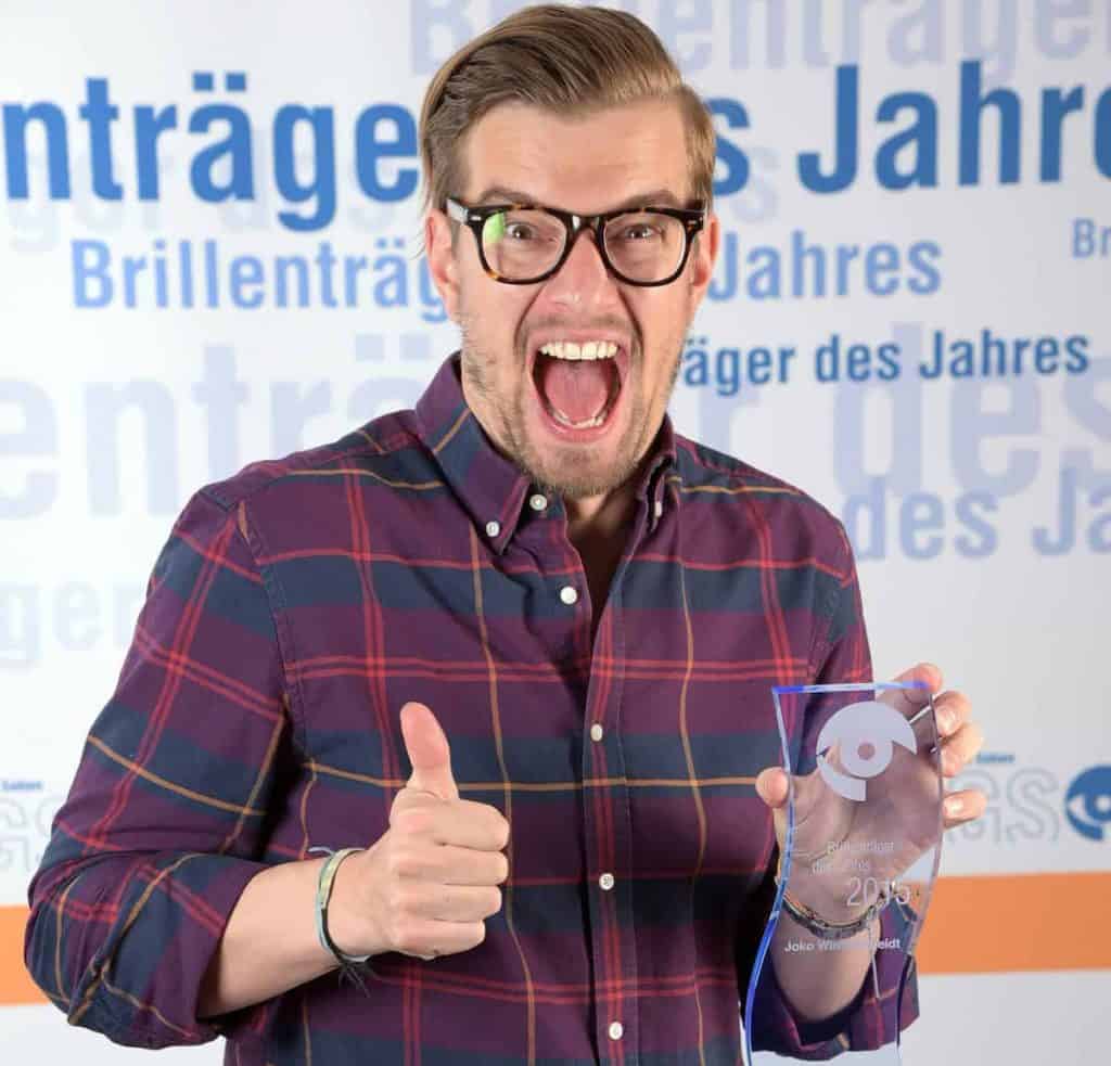 Ein Mann mit Brille und kariertem Hemd lächelt begeistert, streckt den Daumen hoch und hält dabei eine Trophäe aus klarem Glas in der Hand. Im Hintergrund ist ein Text auf Deutsch zu sehen.