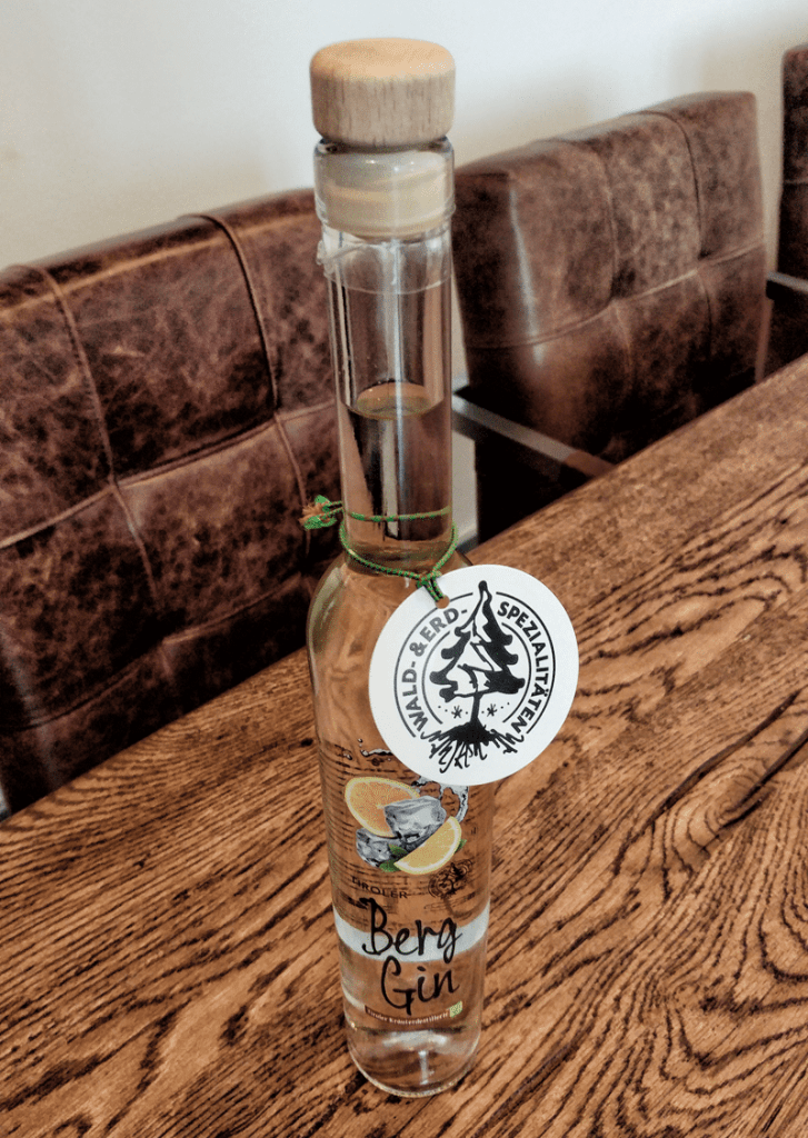Auf einem Holztisch steht eine hohe Glasflasche mit der Aufschrift „Berg Gin“ und einem Holzverschluss. Um den Flaschenhals hängt ein rundes Etikett mit einem Berg-Logo. Im Hintergrund stehen braune Ledersessel.