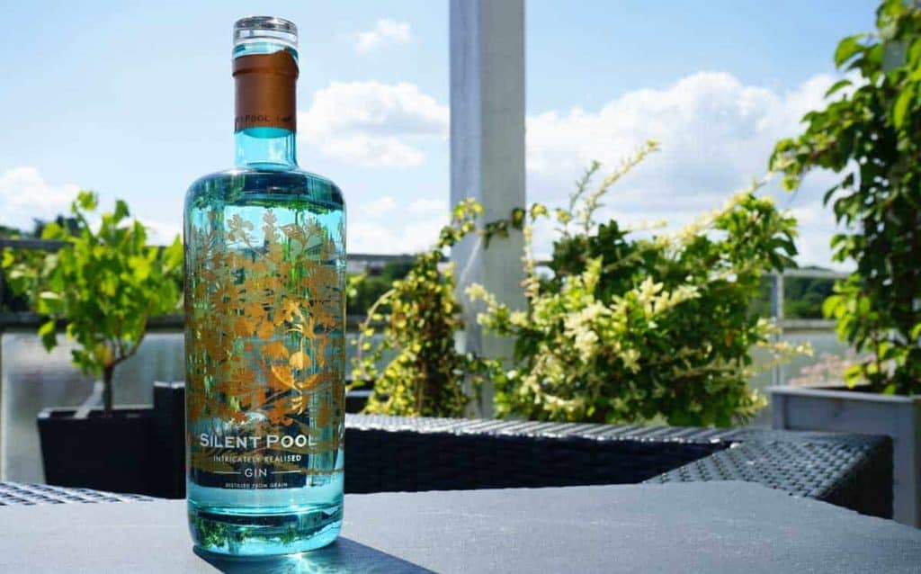 Auf einem Tisch im Freien steht eine Flasche „Silent Pool“-Gin mit floralen Goldverzierungen, umgeben von üppigen grünen Pflanzen und strahlendem Sonnenlicht unter einem blauen Himmel mit flauschigen Wolken.