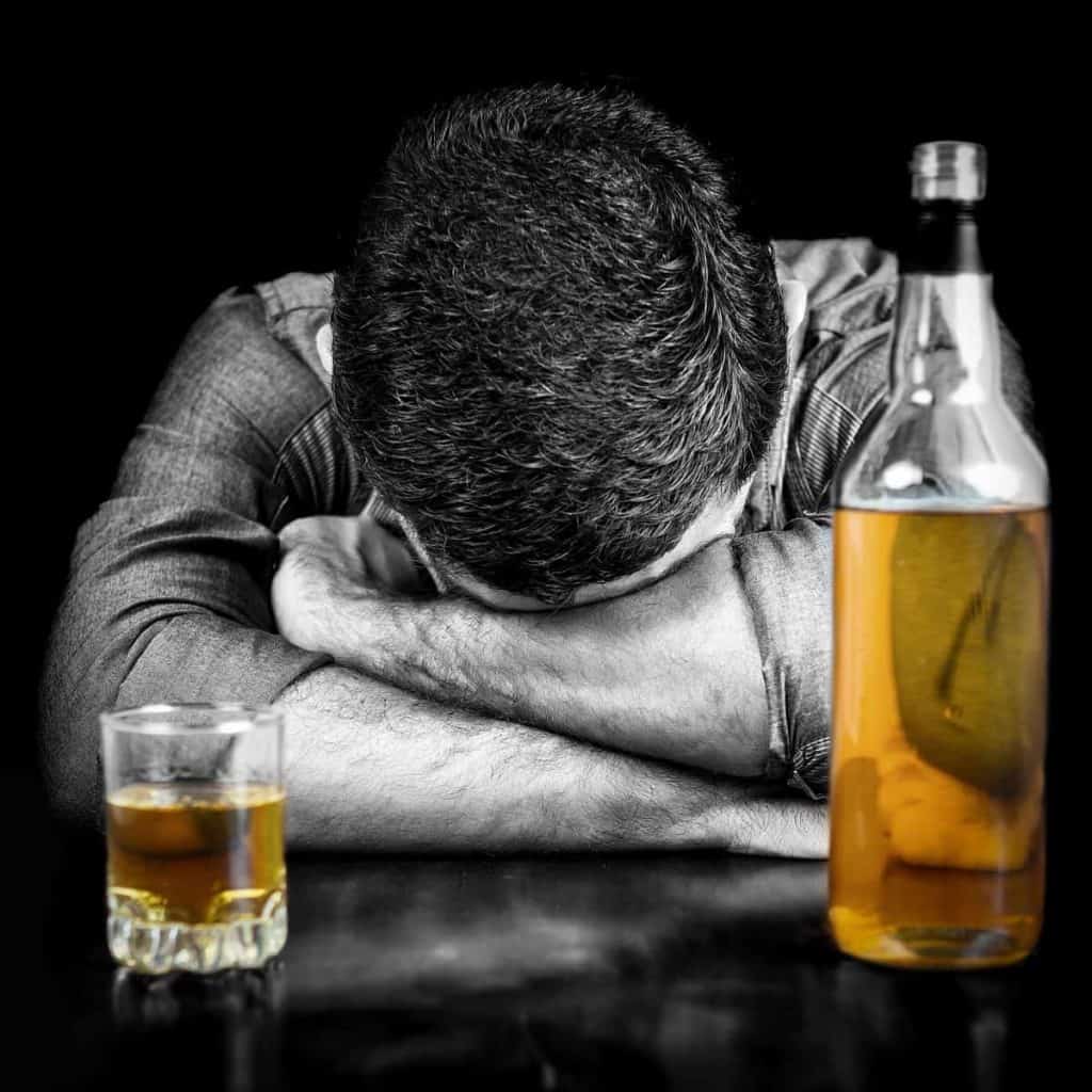 Ein Mann, der den Kopf auf seine verschränkten Arme gestützt hat, sitzt an einem Tisch mit einem Glas Whisky und einer fast vollen Whiskyflasche.