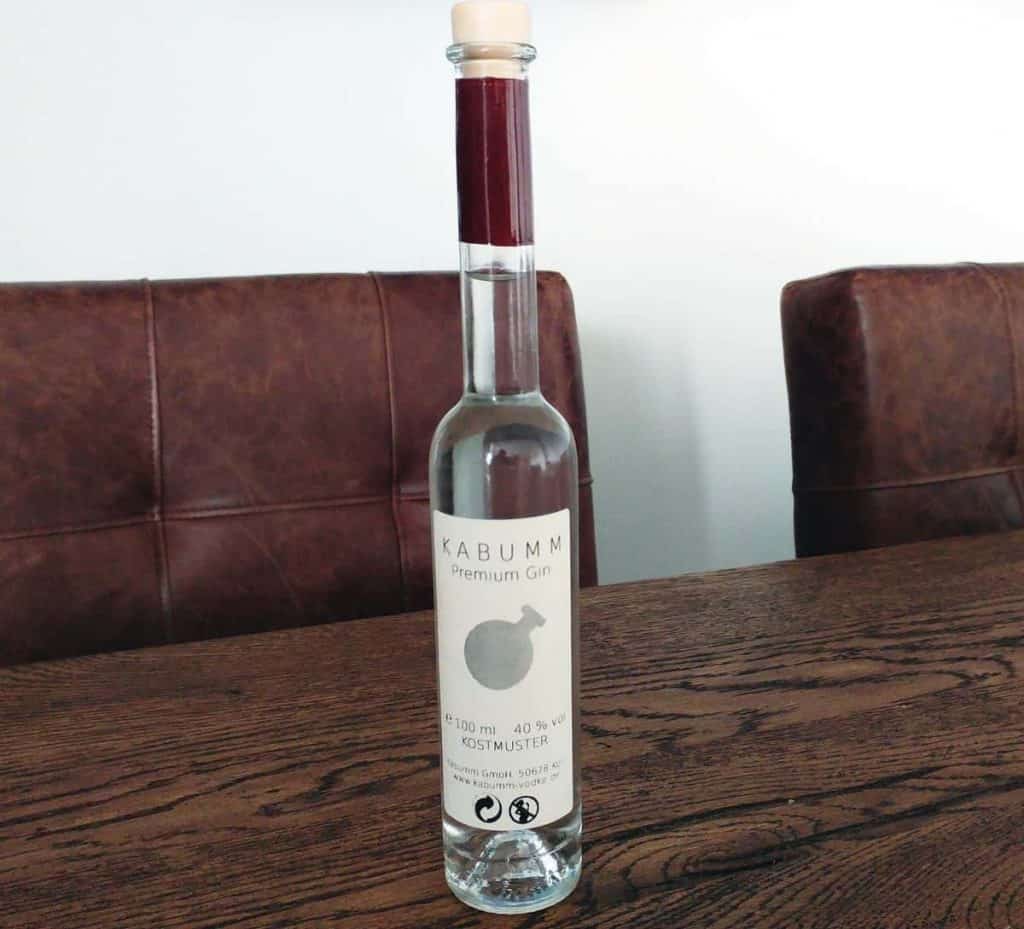 Auf einem Holztisch zwischen zwei braunen Ledersesseln steht eine hohe, durchsichtige Glasflasche mit der Aufschrift „KABUMM Premium Gin“. Die Flasche hat einen Holzkorken und ein schlichtes, minimalistisches Etikettendesign.