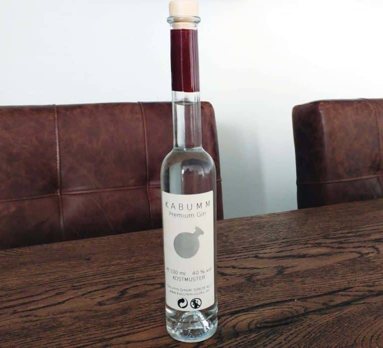 Auf einem Holztisch zwischen zwei braunen Ledersesseln steht eine hohe, durchsichtige Glasflasche mit der Aufschrift „KABUMM Premium Gin“. Die Flasche hat einen Holzkorken und ein schlichtes, minimalistisches Etikettendesign.