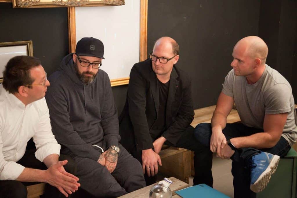 Vier Männer sitzen eng beieinander und unterhalten sich; einer gestikuliert beim Sprechen, zwei hören aufmerksam zu, und einer hat die Hände gefaltet. Sie befinden sich in einem Raum, im Hintergrund sind Kunstwerke zu sehen.