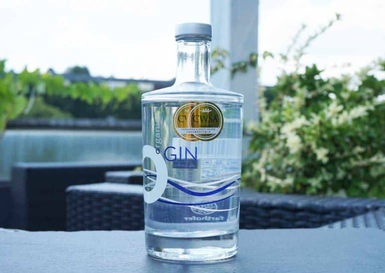 Auf einem Tisch im Freien steht eine klare Glasflasche mit Bio-Gin, auf deren Etikett eine Goldauszeichnung zu sehen ist; im Hintergrund sind Grünpflanzen, ein Gewässer und Terrassenmöbel zu sehen.