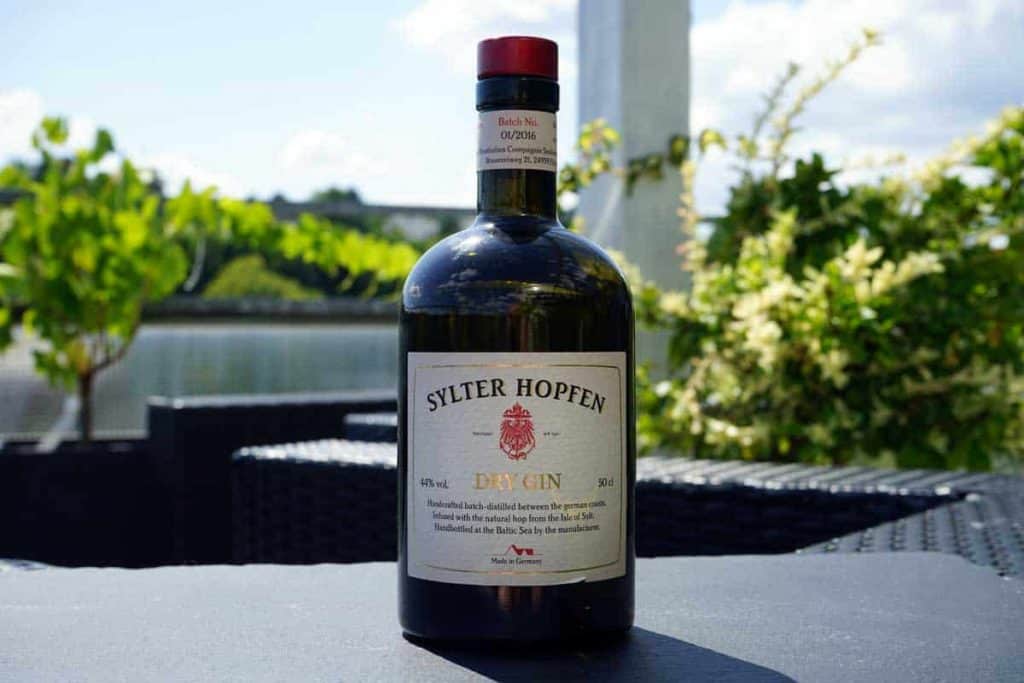 Auf einem Tisch im Freien steht eine Flasche „Sylter Hopfen Dry Gin“, im unscharfen Hintergrund sind an einem sonnigen Tag Grünflächen und ein Gewässer zu sehen.