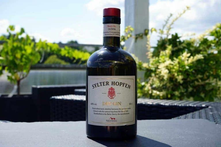 Auf einem Tisch im Freien steht eine Flasche „Sylter Hopfen Dry Gin“, im unscharfen Hintergrund sind an einem sonnigen Tag Grünflächen und ein Gewässer zu sehen.