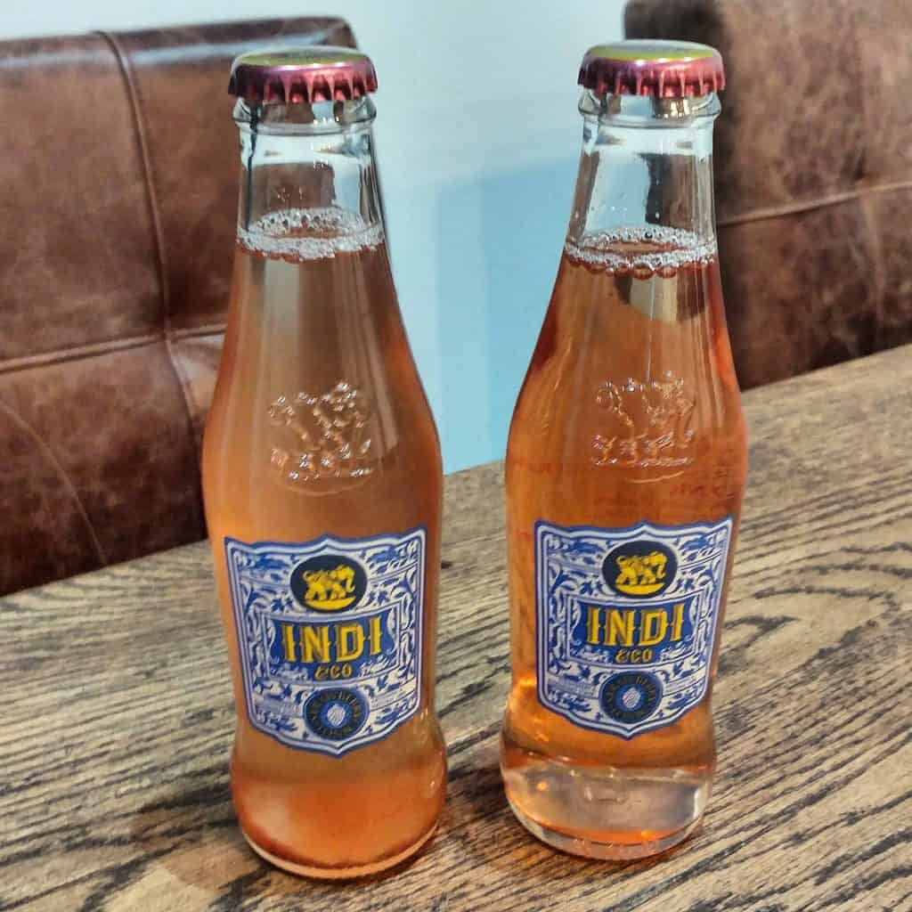 Auf einem Holztisch stehen zwei Glasflaschen mit rosa Limonade, die mit der Aufschrift „Indi“ und kunstvollen blauen und gelben Mustern verziert sind; im Hintergrund sind braune Ledersessel zu sehen.