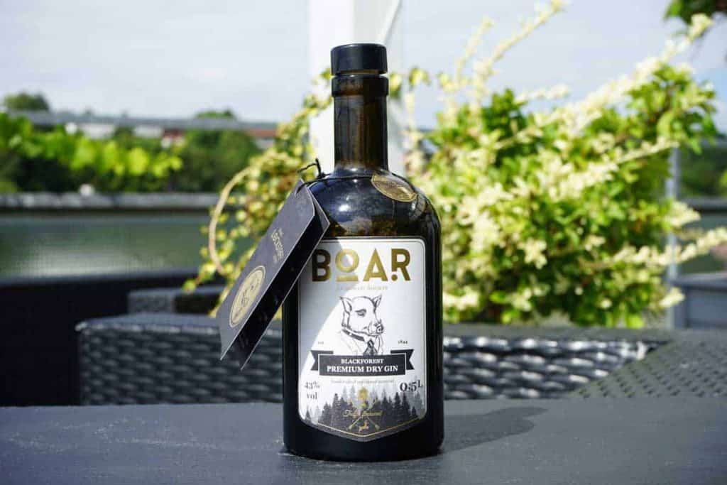 Auf einem Tisch im Freien steht eine Flasche „BOAR Premium Dry Gin“ mit einem schwarz-weißen Etikett, auf dem ein Wildschwein abgebildet ist; im unscharfen Hintergrund sind Grünpflanzen und Wasser zu sehen.