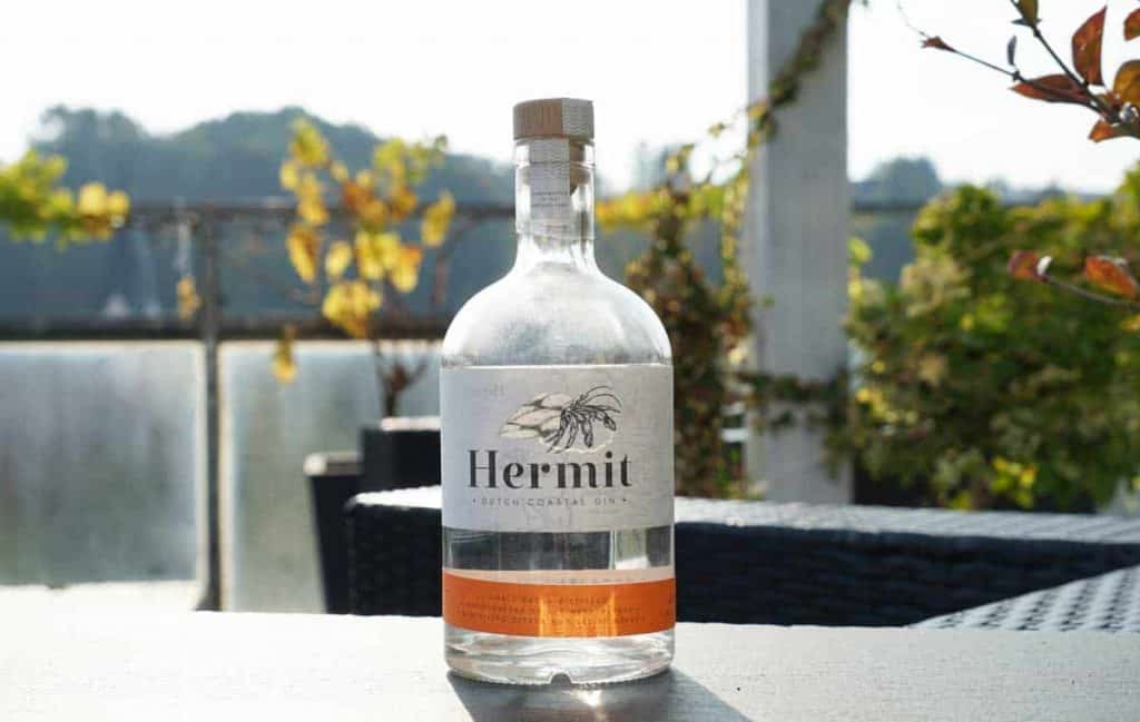 Auf einem Tisch im Freien steht eine durchsichtige Flasche „Hermit Dutch Coastal Gin“, im unscharfen Hintergrund sind Terrassenmöbel und Pflanzen zu sehen.