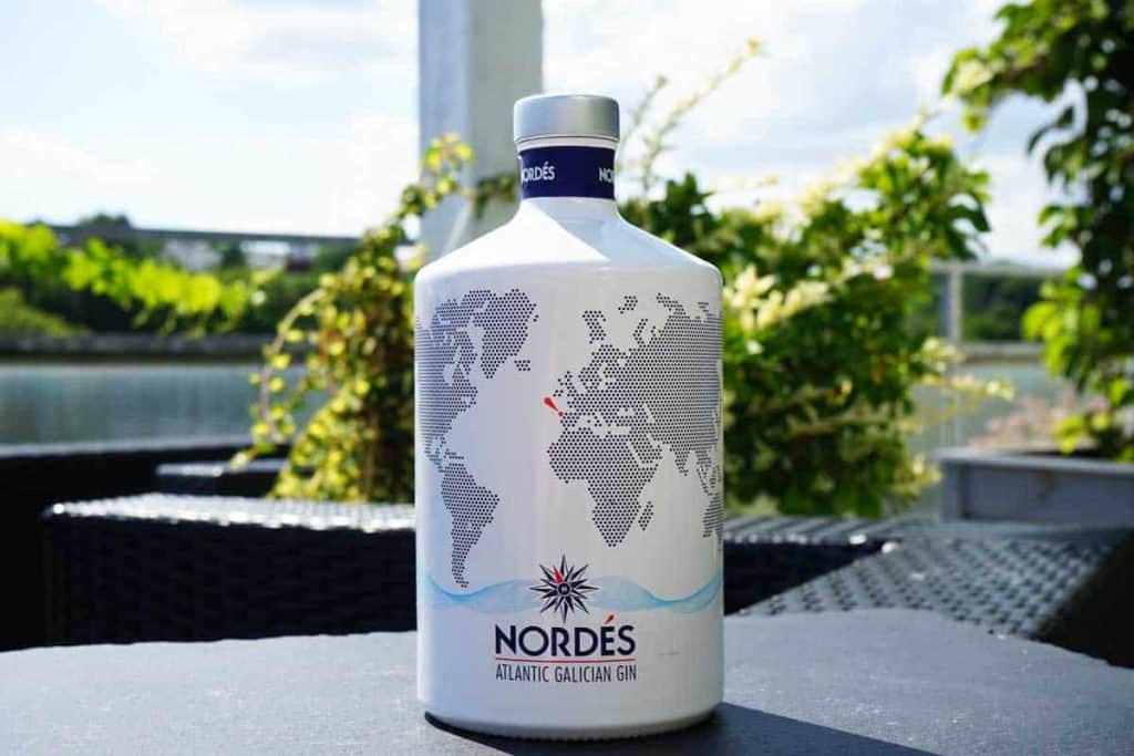 Eine Flasche „Nordés Atlantic Galician Gin“ mit einem Weltkarten-Motiv steht an einem Tisch im Freien, im Hintergrund sind an einem sonnigen Tag Grünflächen und ein Gewässer zu sehen.