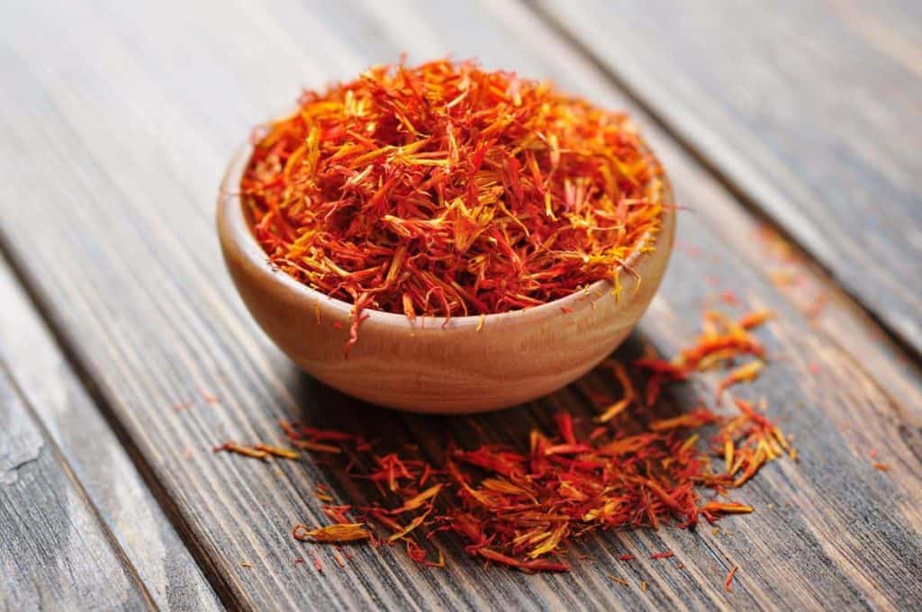 Eine kleine Holzschale, gefüllt mit leuchtend orangefarbenen und roten Safranfäden, von denen einige auf einer rustikalen Holzoberfläche verstreut liegen.