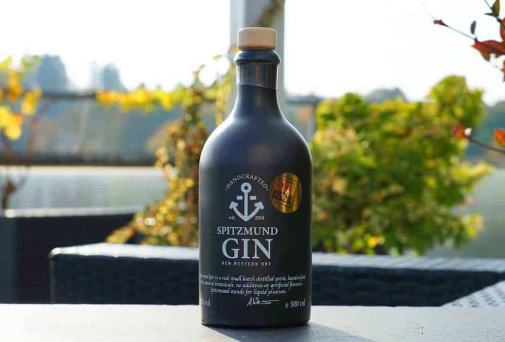 Auf einem Tisch im Freien steht eine schwarze Flasche Spitzmund Gin, im Hintergrund sind verschwommene grüne Blätter und Sonnenlicht zu sehen. Die Flasche hat ein goldenes Siegel und weißen Text auf dem Etikett.