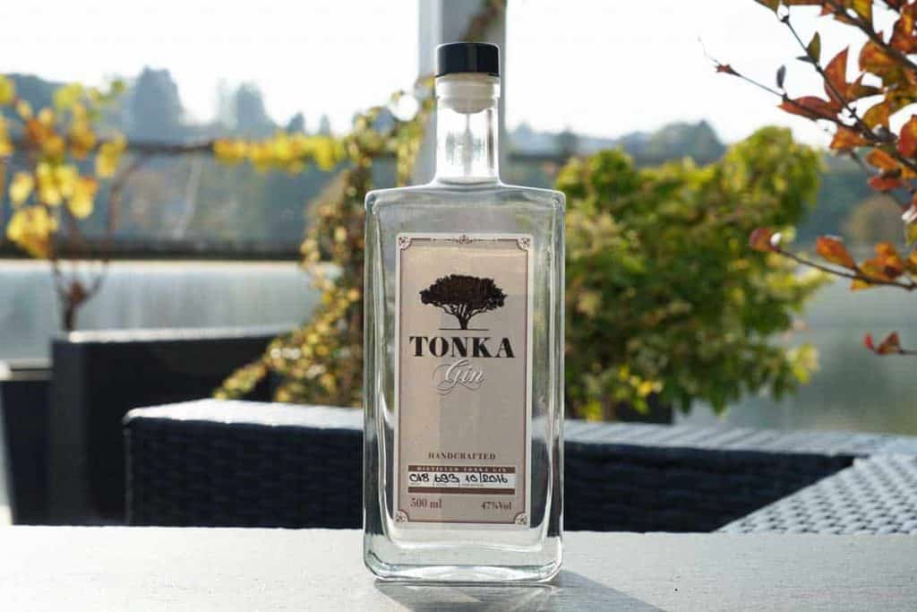 Auf einem Tisch im Freien steht eine klare Glasflasche Tonka Gin, im Hintergrund sind Pflanzen, Laub und Gartenmöbel im natürlichen Licht zu sehen, wobei der Hintergrund unscharf wirkt.