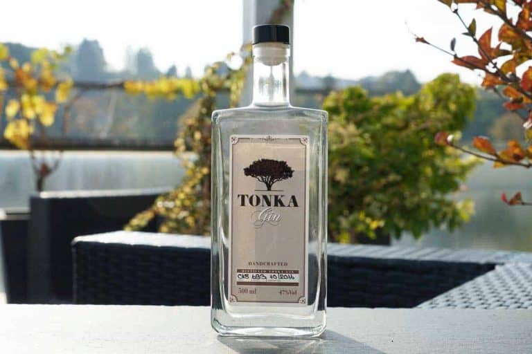 Auf einem Tisch im Freien steht eine klare Glasflasche Tonka Gin, im Hintergrund sind Pflanzen, Laub und Gartenmöbel im natürlichen Licht zu sehen, wobei der Hintergrund unscharf wirkt.