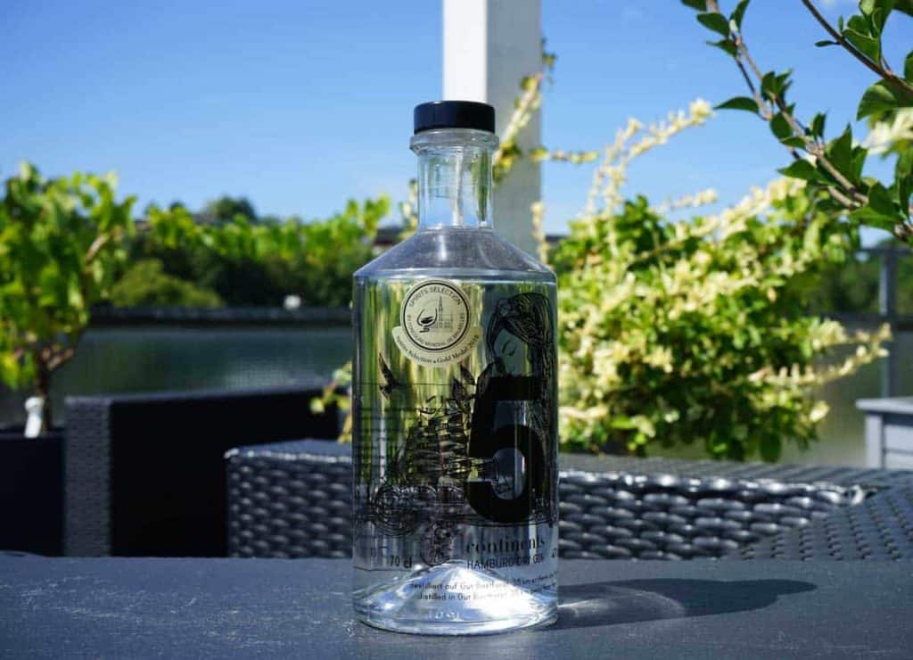 Auf einem Tisch im Freien steht eine durchsichtige Flasche mit der Aufschrift „5“, im Hintergrund sind grüne Pflanzen, ein See und ein blauer Himmel zu sehen. Die Flasche ist mit schwarzen Illustrationen verziert und weist am Flaschenhals ein goldenes Medaillon auf.