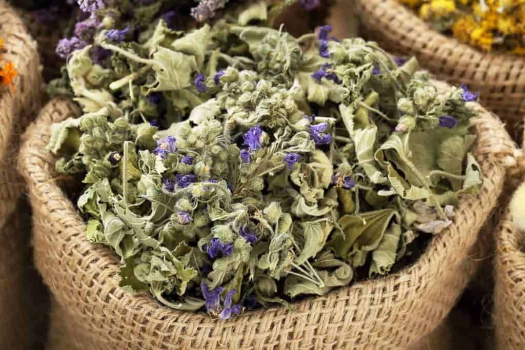 Ein Jutesack, gefüllt mit getrockneten Kräutern und kleinen violetten Blüten; im Hintergrund sind weitere Säcke mit getrockneten Pflanzen zu sehen.
