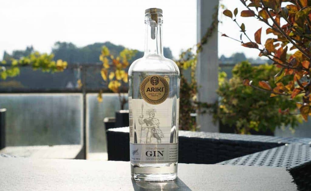 Auf einem Tisch im Freien steht eine durchsichtige Flasche Ariki-Gin, durch die das Sonnenlicht scheint; im Hintergrund ist Laub zu sehen, und daneben stehen Terrassenmöbel.