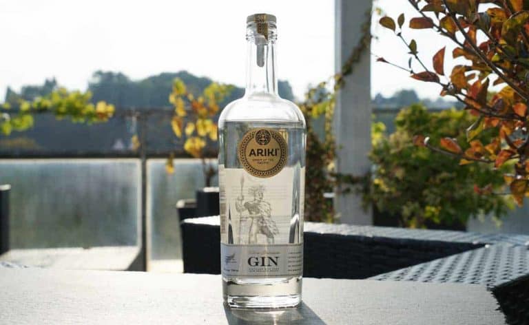 Auf einem Tisch im Freien steht eine durchsichtige Flasche Ariki-Gin, durch die das Sonnenlicht scheint; im Hintergrund ist Laub zu sehen, und daneben stehen Terrassenmöbel.