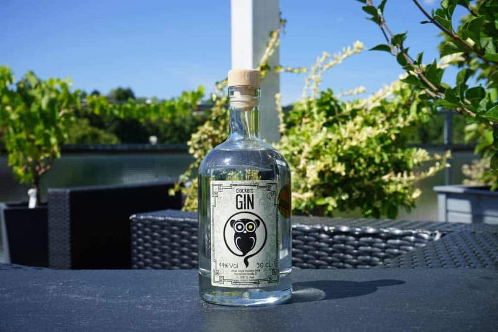 Auf einem Tisch im Freien steht eine Gin-Flasche aus klarem Glas mit einem schwarz-weißen Eulenlogo, umgeben von grünen Pflanzen und unter einem strahlend blauen Himmel.