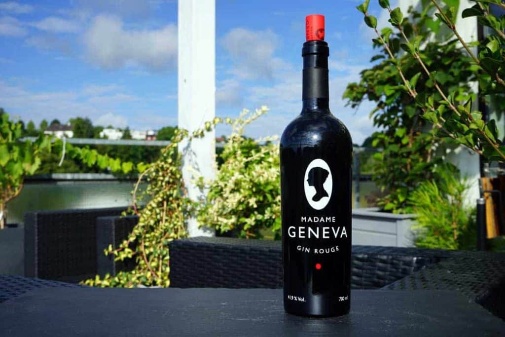 Auf einem Tisch im Freien steht eine schwarze Flasche „Madame Geneva Gin Rouge“, im Hintergrund sind Grünpflanzen und ein klarer Himmel zu sehen.