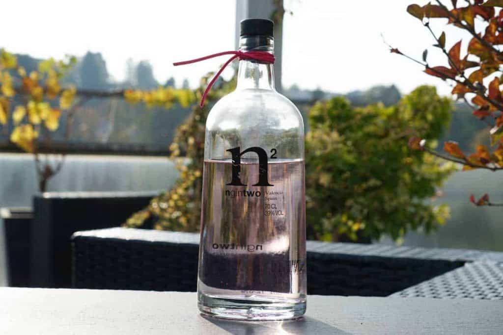 Auf einem Tisch steht eine klare Glasflasche mit einem schwarzen Verschluss und einer roten Schleife. Die Flasche trägt die Aufschrift „n2“ und ist mit einer durchsichtigen Flüssigkeit gefüllt. Im Hintergrund sind Grünpflanzen im Freien und Sonnenlicht zu sehen.