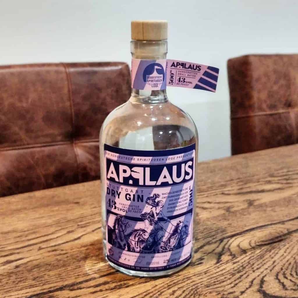 Auf einem Holztisch steht eine Flasche „Applaus Dry Gin“ mit einem violett-weißen Etikett. Im Hintergrund stehen zwei braune Polstersessel.