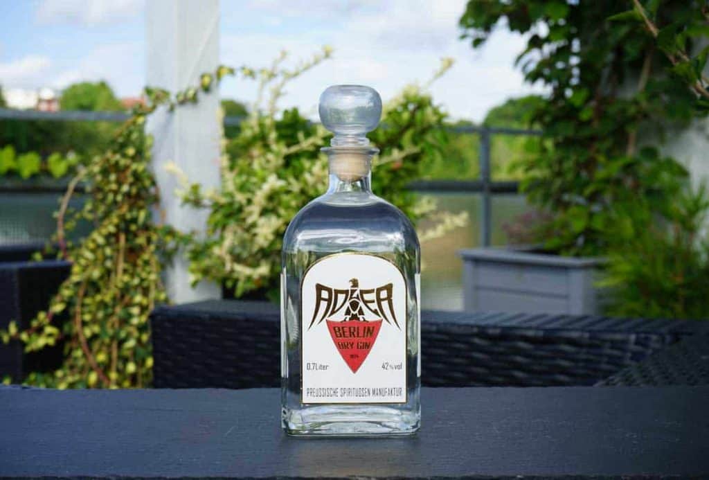 Auf einem dunklen Tisch im Freien steht eine klare Glasflasche „Adaka Berlin Pink Gin“, im Hintergrund sind Pflanzen und grünes Laub verschwommen zu erkennen.