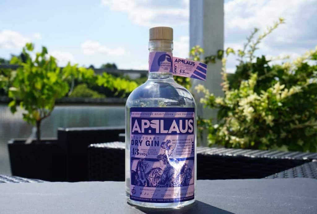 Auf einem schwarzen Gartentisch steht eine Flasche APPLAUS Dry Gin mit violettem Etikett, im Hintergrund sind grüne Pflanzen und ein blauer Himmel zu sehen.
