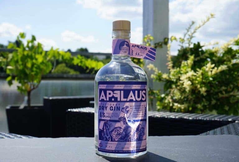 Auf einem schwarzen Gartentisch steht eine Flasche APPLAUS Dry Gin mit violettem Etikett, im Hintergrund sind grüne Pflanzen und ein blauer Himmel zu sehen.