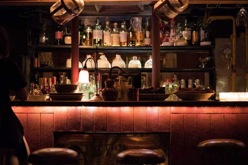 Eine schwach beleuchtete Bar mit Regalen voller verschiedener Spirituosenflaschen hinter der Theke, warmer Beleuchtung, zwei Barhockern davor und verschiedenen Gläsern und Dekorationsgegenständen auf der Theke.