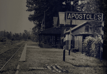 Ein Schwarz-Weiß-Foto eines ländlichen Bahnhofs mit einem Schild, auf dem „APOSTOLES“ steht. Der Bahnsteig verläuft parallel zu den Gleisen und ist von Bäumen und alten Gebäuden umgeben. Die Szene wirkt ruhig und verlassen.