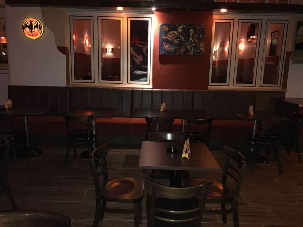 Ein schwach beleuchtetes Restaurant mit leeren Stühlen und Tischen aus dunklem Holz, braunen gepolsterten Sitznischen, Wandbildern und einem leuchtenden Bacardi-Fledermaus-Logo in der oberen linken Ecke.
