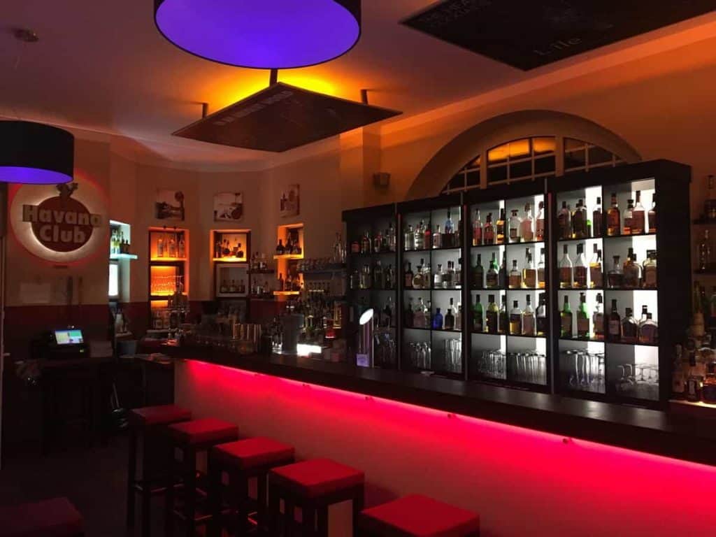 Eine moderne Bar mit einer rot beleuchteten Theke, Regalen voller Flaschen, hohen Barhockern und einer stimmungsvollen Beleuchtung in Lila und Gelb. An der Wand ist ein „Havana Club“-Schild zu sehen.