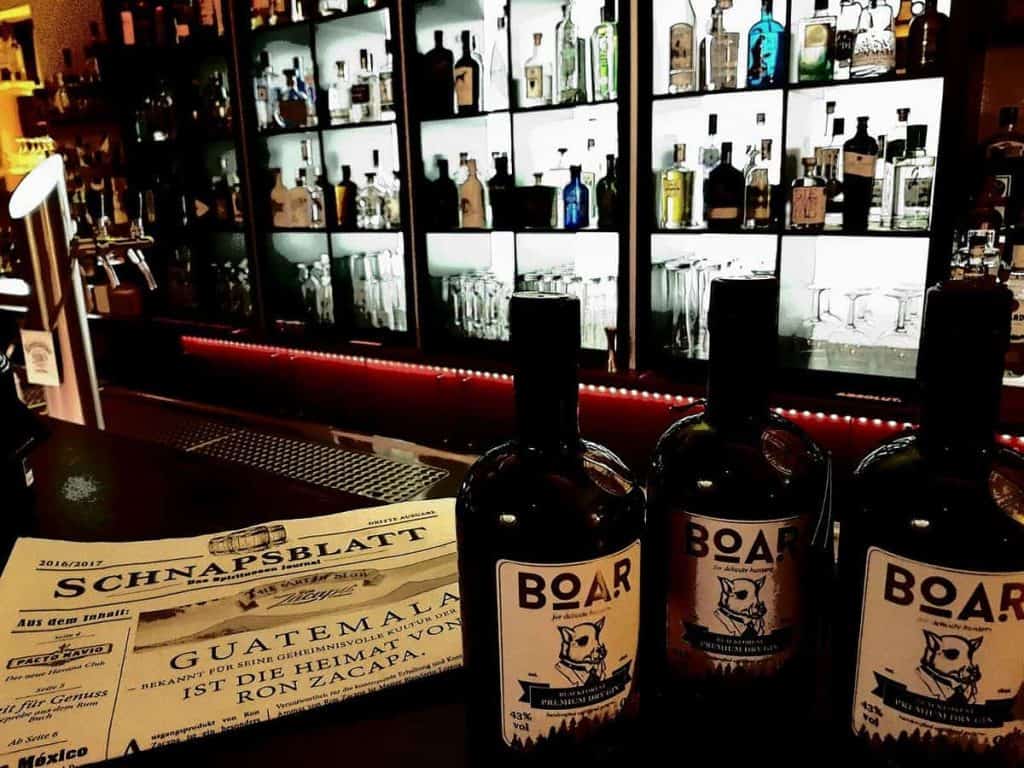 Drei Flaschen mit der Aufschrift „BOAR“ stehen auf einer Bartheke neben einer Speisekarte mit dem Titel „Schnapsblatt“, während im Hintergrund Regale mit verschiedenen Spirituosenflaschen und Gläsern beleuchtet sind.