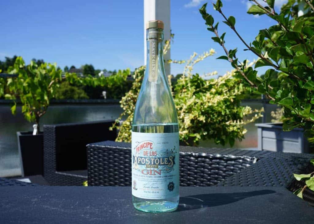 Auf einem dunklen Tisch im Freien, umgeben von grünen Pflanzen und Wasser, steht im sonnigen Hintergrund eine Flasche Gin mit der Aufschrift „Principe de los Apostoles“.