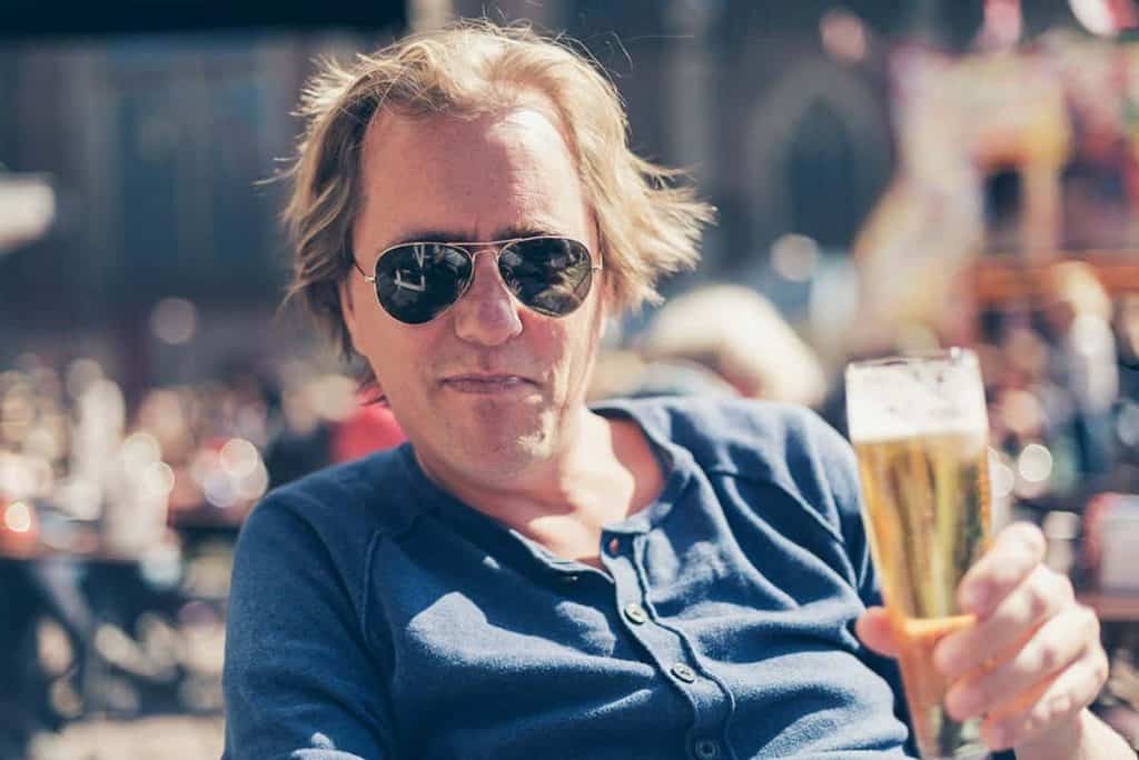 Ein Mann mit Sonnenbrille und blauem Hemd sitzt im Freien und hält ein Glas Bier in der Hand. Der Hintergrund ist unscharf und zeigt Menschen sowie eine farbenfrohe Umgebung, was auf einen lebhaften, sonnigen Tag hindeutet.