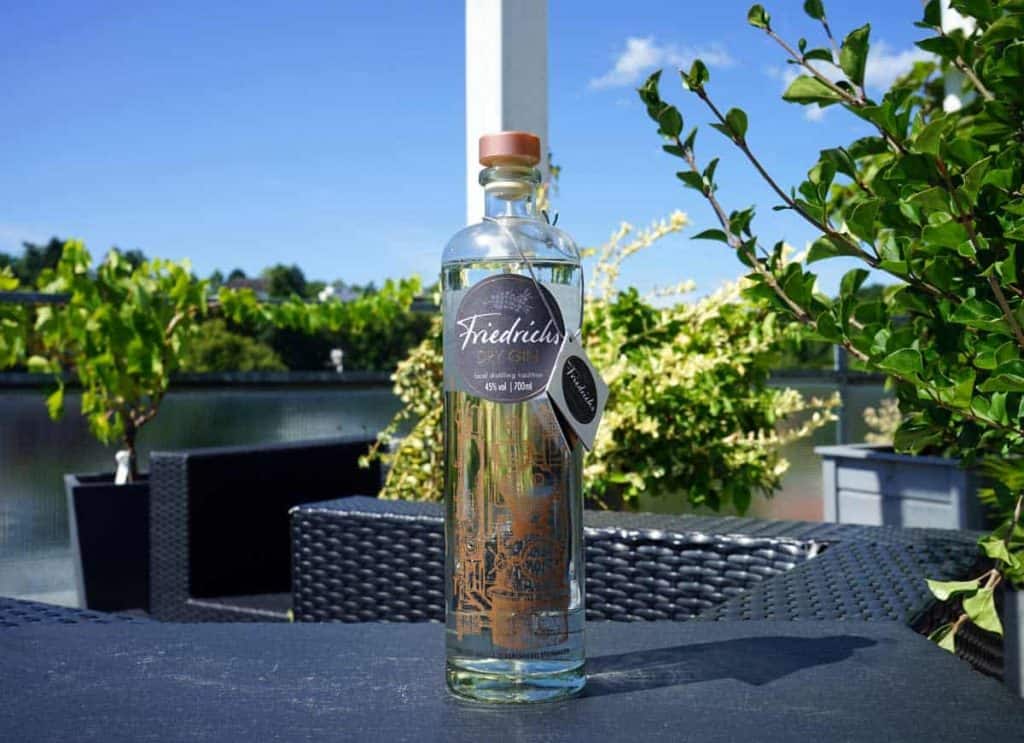 Auf einem dunklen Tisch im Freien, umgeben von grünen Pflanzen und Korbstühlen, steht eine Flasche Friedrichs Dry Gin, im Hintergrund ist unter blauem Himmel ein ruhiges Gewässer zu sehen.