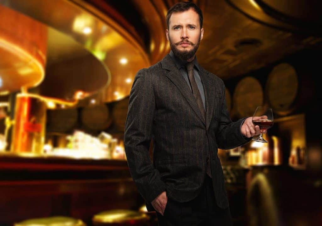 Ein Mann in einem dunklen Anzug, der ein Glas Brandy in der Hand hält, steht selbstbewusst in einer schummrig beleuchteten Bar, deren Interieur in Goldtönen gehalten ist und in deren Hintergrund Fässer zu sehen sind.