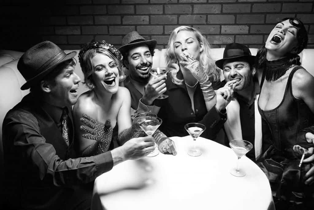 Sechs Personen in Retro-Kleidung, die lachend an einem kleinen Tisch in einer lebhaften, festlichen Atmosphäre Cocktails genießen. Das Foto ist in Schwarz-Weiß gehalten und vermittelt das Flair der 1920er Jahre oder einer „Speakeasy“-Bar.