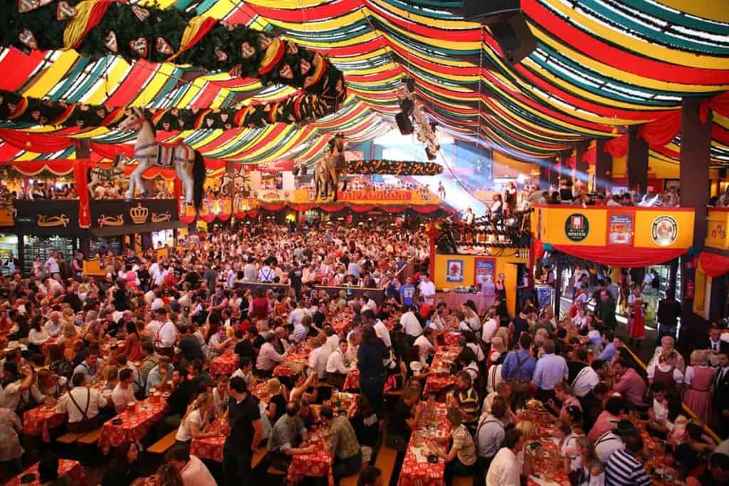 Ein großer, gut besuchter Bierkeller, geschmückt mit bunten Transparenten und Fahnen. Die Gäste sitzen an langen Tischen, essen, trinken und unterhalten sich. Die festliche Atmosphäre ist lebhaft und fröhlich, über den Köpfen hängen Fässer und Pferdedekorationen.