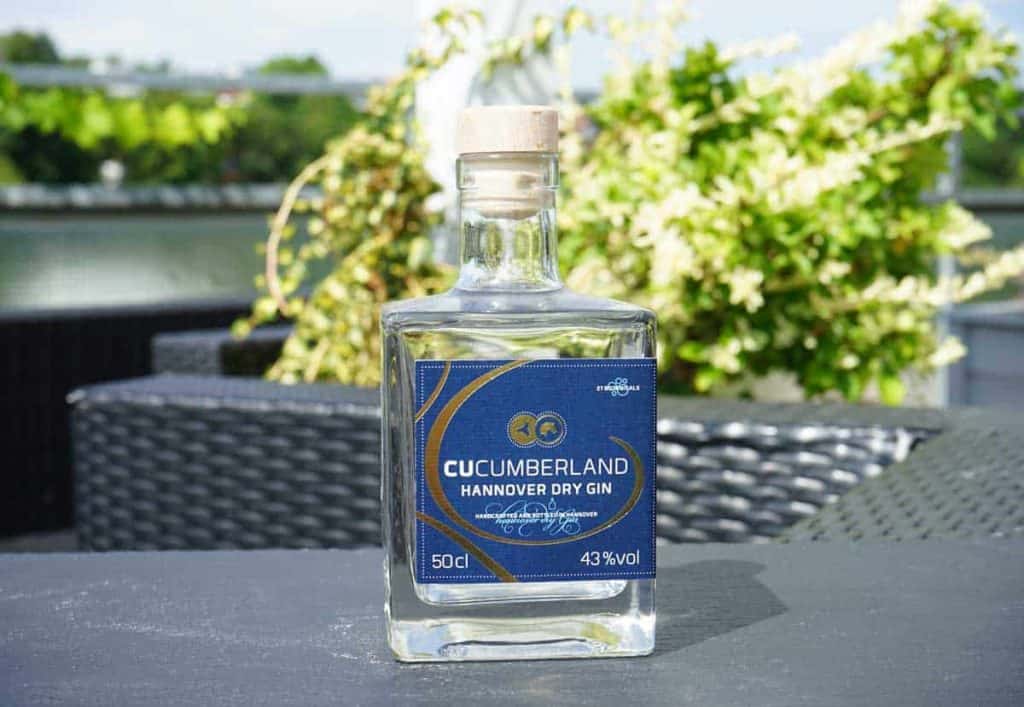 Auf einem Tisch im Freien steht eine quadratische Glasflasche „Cumberland Hannover Dry Gin“, im Hintergrund sind Grünpflanzen und Korbmöbel zu sehen.