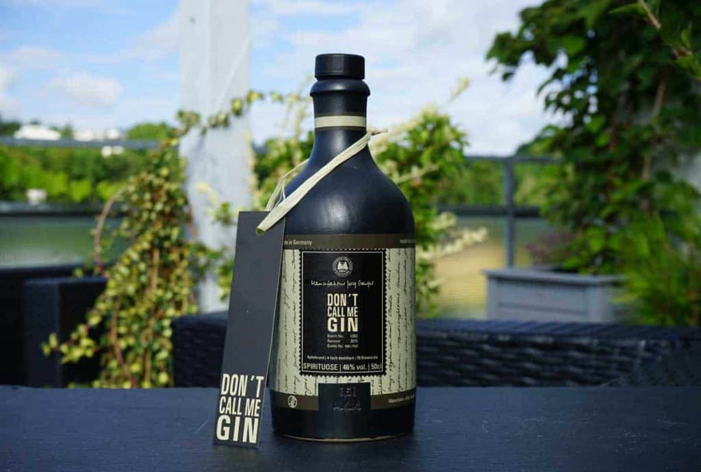 Auf einem dunklen Tisch im Freien, umgeben von Grünpflanzen und vor einem unscharfen Hintergrund, steht eine schwarze Flasche mit der Aufschrift „Dont Call Me Gin“. Am Flaschenhals hängt ein passendes Etikett.