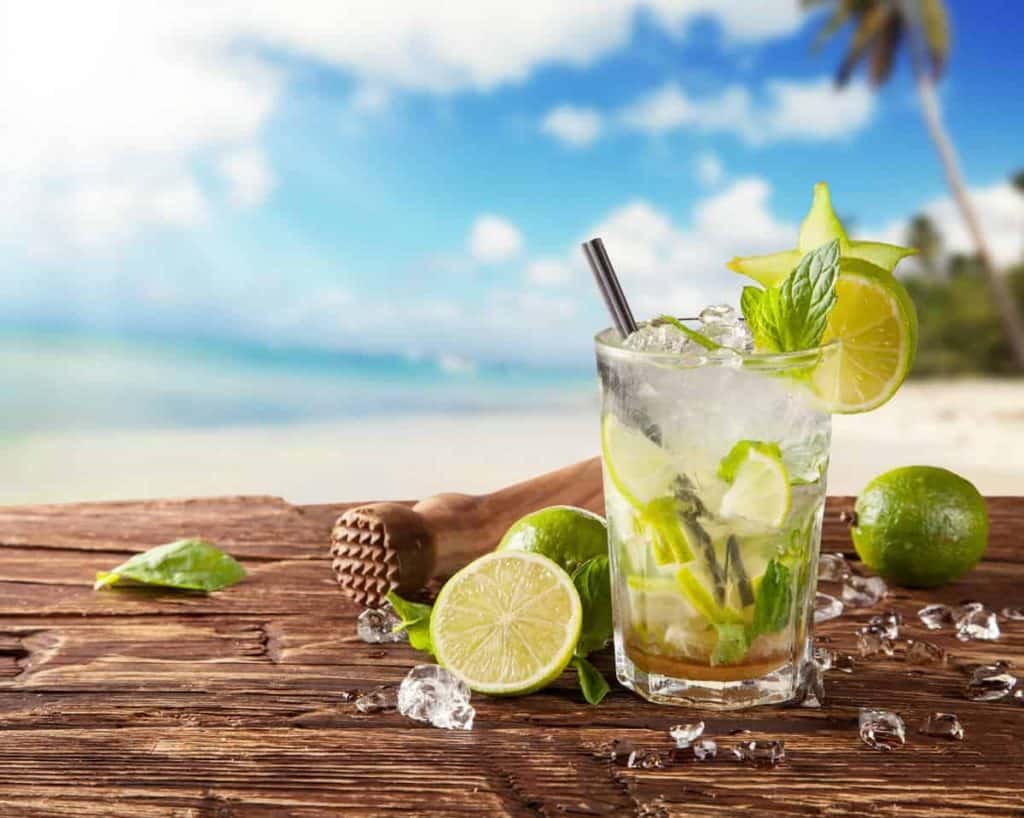 Auf einem Holztisch am Strand steht ein Mojito-Cocktail mit Limette, Minze, Eis und einem Strohhalm; daneben liegen Limetten, Minzblätter, Eiswürfel und ein Stößel. Im Hintergrund sind Sand, Meer und Palmen zu sehen.
