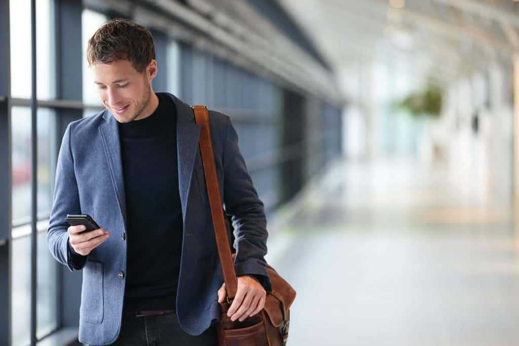 Ein Mann in einem blauen Blazer mit einer braunen Umhängetasche lächelt, während er auf sein Handy schaut; er steht in einem hellen, modernen Flur mit großen Fenstern.