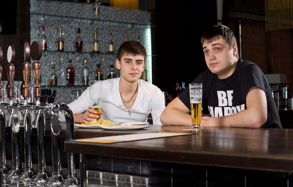 Zwei junge Männer sitzen mit Getränken und einem Teller Essen an einer Bar und blicken in die Kamera. Im Hintergrund stehen Bierhähne und Flaschen in Regalen.