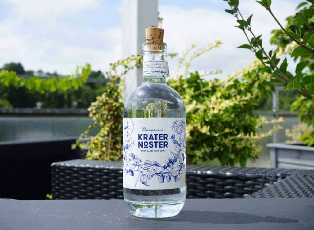 Eine durchsichtige Glasflasche mit der Aufschrift „Krater Noster“ und einem Korken steht auf einem Tisch im Freien, umgeben von Grünpflanzen und Terrassenmöbeln, im Hintergrund sind Wasser und Bäume zu sehen.