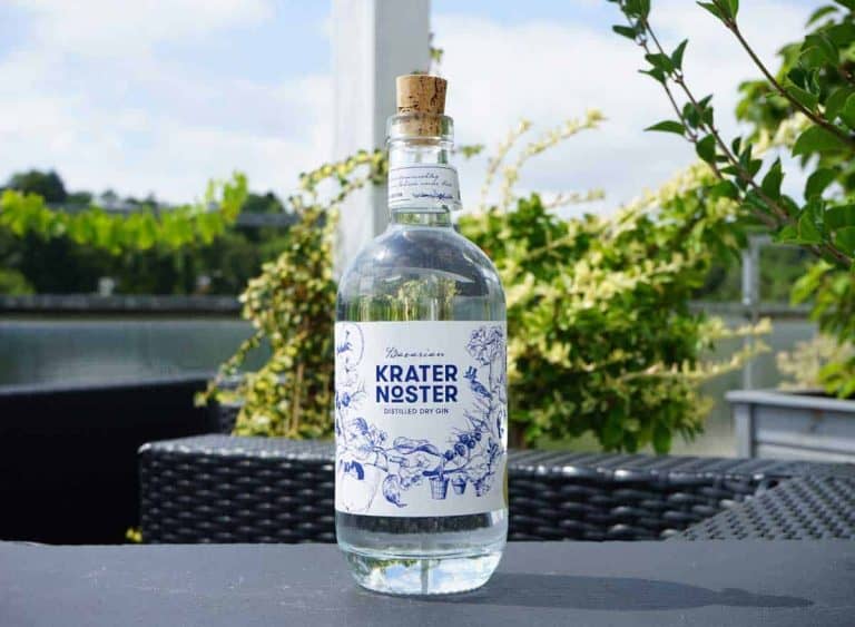 Eine durchsichtige Glasflasche mit der Aufschrift „Krater Noster“ und einem Korken steht auf einem Tisch im Freien, umgeben von Grünpflanzen und Terrassenmöbeln, im Hintergrund sind Wasser und Bäume zu sehen.