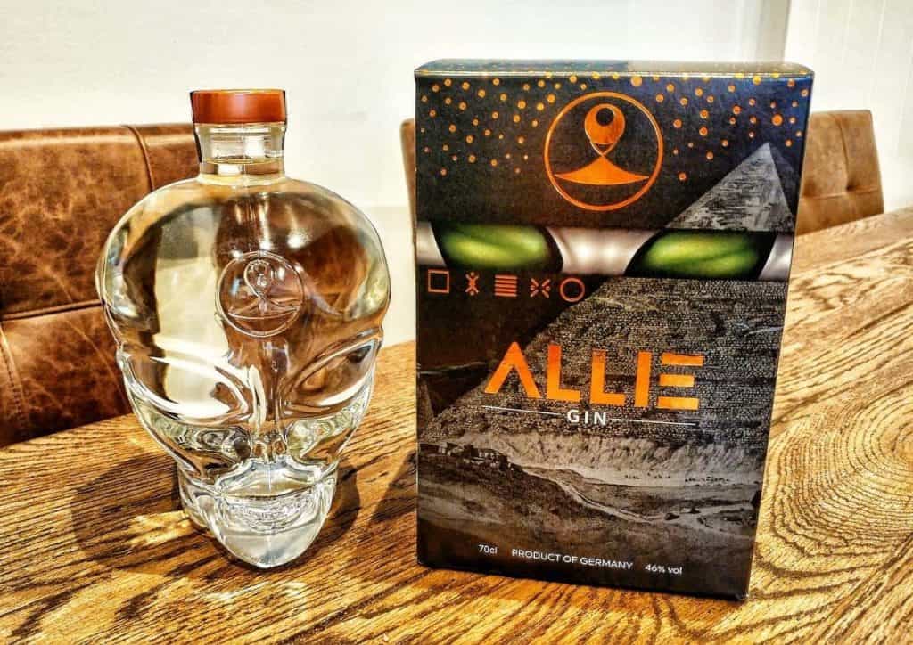 Eine glasene Flasche in Form eines Schädels neben einer Schachtel mit der Aufschrift „ALLIE GIN“, verziert mit grünen Alien-Augen und Pyramidenmotiven, auf einem Holztisch, im Hintergrund braune Ledersessel.
