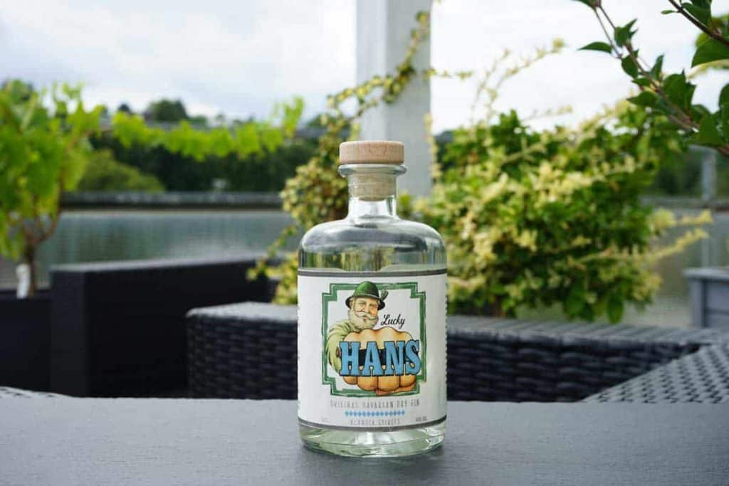 Auf einem dunklen Tisch im Freien steht eine durchsichtige Flasche mit der Aufschrift „Lady Hans“, im unscharfen Hintergrund sind grünes Laub und Terrassenmöbel zu sehen.