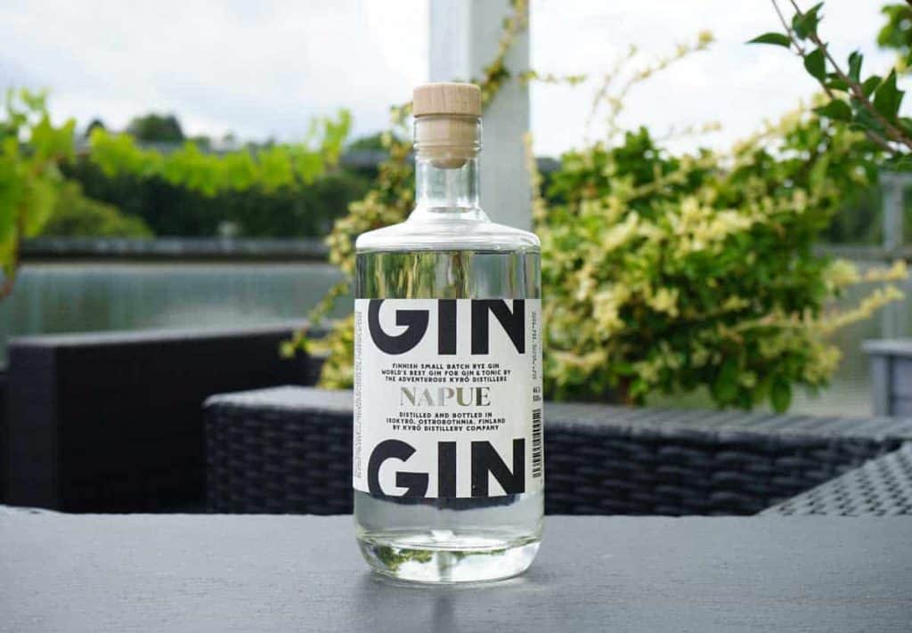 Auf einem dunklen Tisch im Freien steht eine Flasche Napue-Gin mit einem Holzverschluss; im unscharfen Hintergrund sind Korbstühle, Grünpflanzen und Wasser zu sehen.